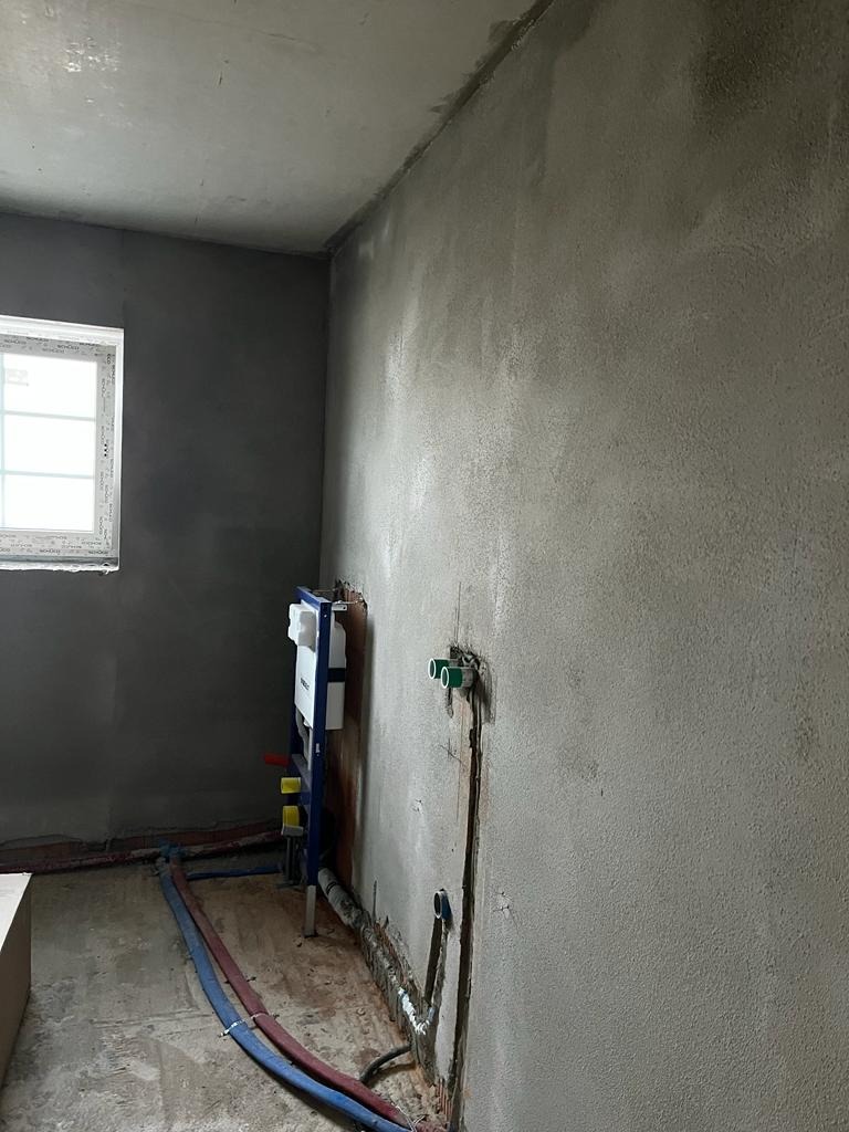 Surowe wnętrze łazienki w trakcie remontu: ściany pokryte świeżym tynkiem, widoczne rury instalacji wodnej i stelaż podtynkowy WC. Okno po lewej stronie kadru.