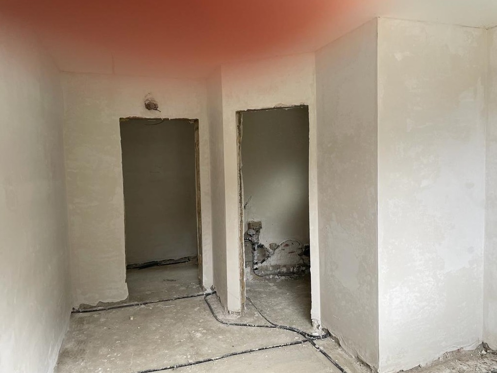 Świeżo otynkowane wnętrze w trakcie remontu, widoczne dwa otwory drzwiowe, szare ściany z nierówną fakturą, kable na podłodze, surowy stan wykończenia.