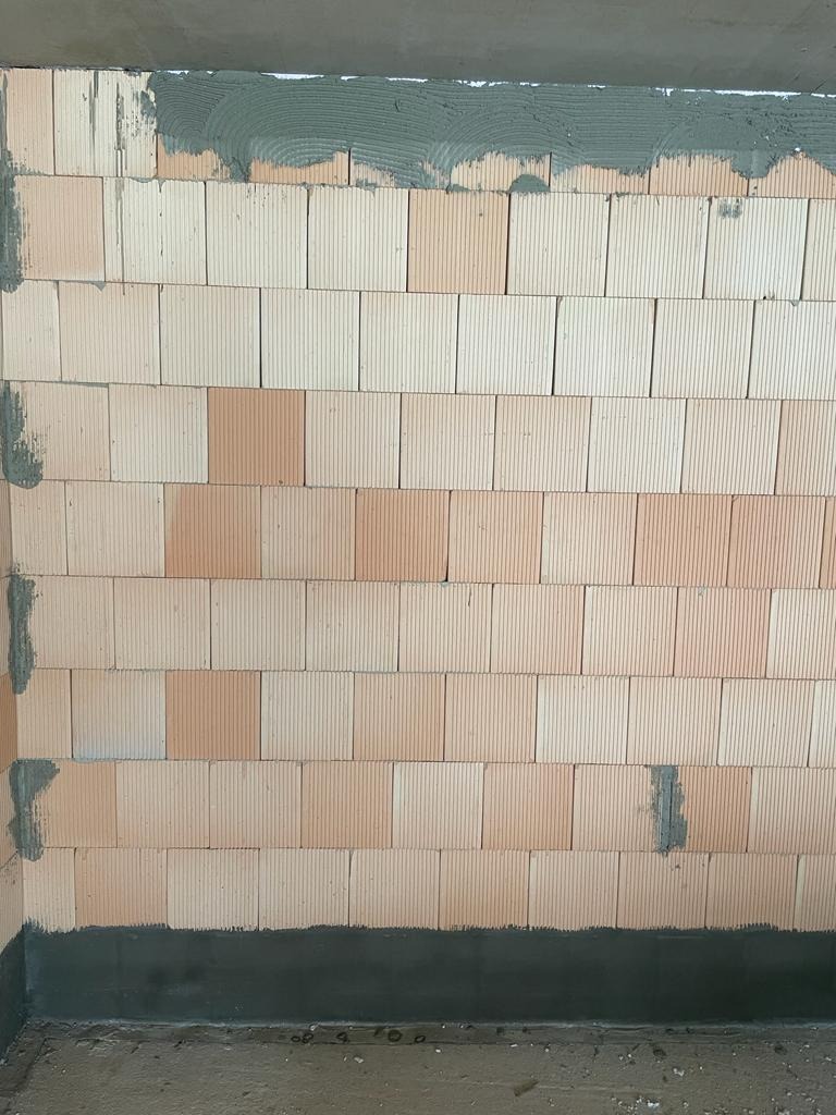 Ściana z pustaków ceramicznych, częściowo pokryta zaprawą, widoczne pionowe żłobienia na pustakach, szara zaprawa na górze i dole ściany.