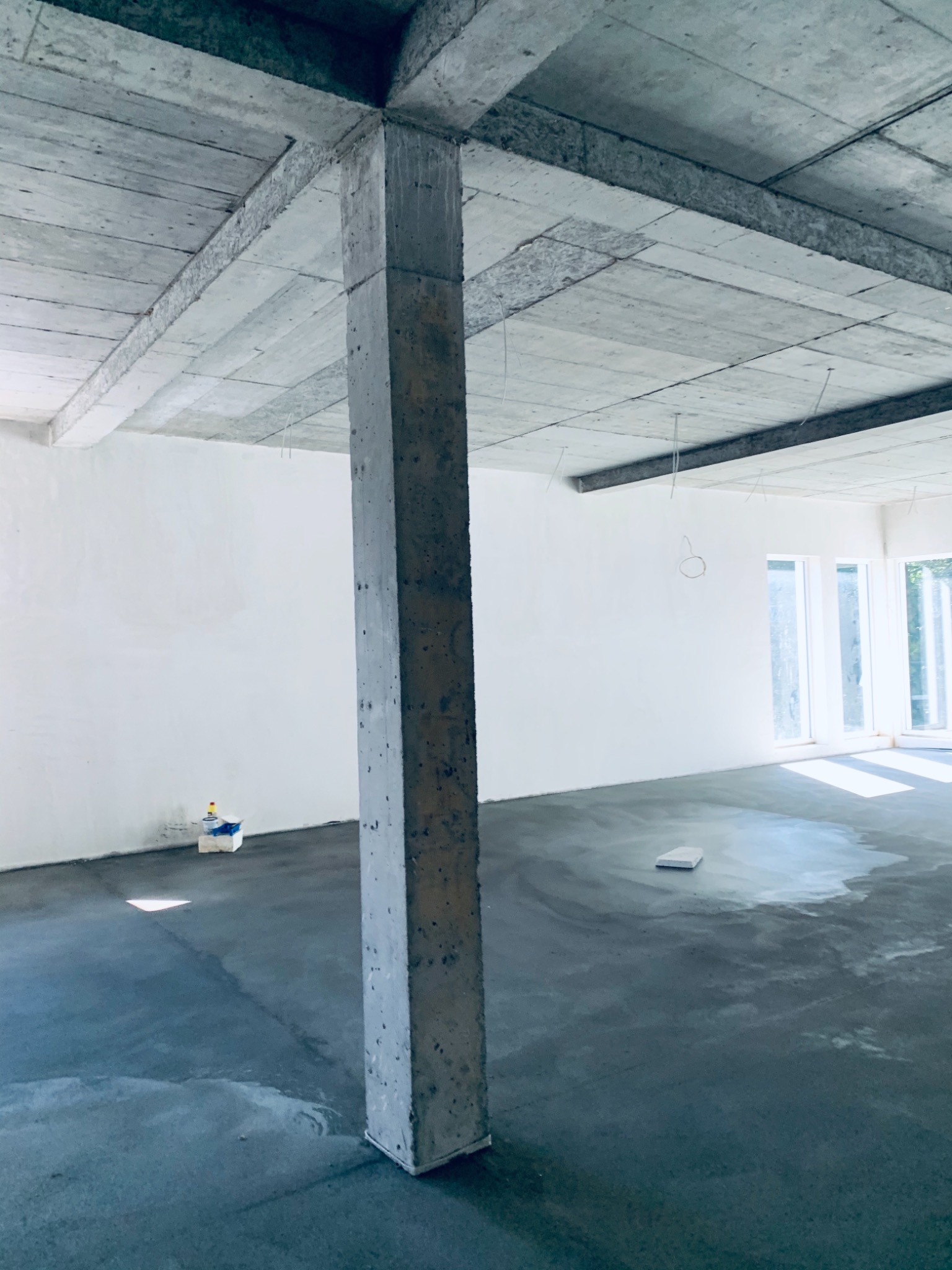 Surowe wnętrze w trakcie prac wykończeniowych: betonowy strop z widocznymi płytami, szary wylewany beton na podłodze, białe ściany i kolumna nośna. W tle okna z widokiem na zieleń.