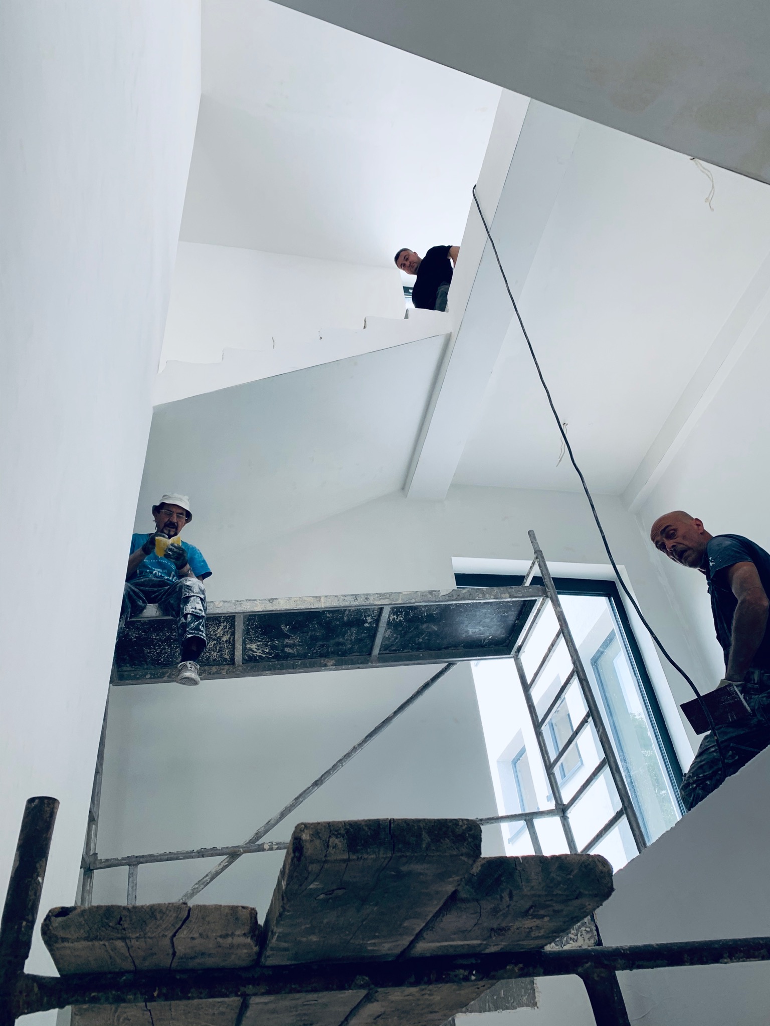 Trzech tynkarzy pracuje w jasnym wnętrzu na różnych poziomach, jeden na rusztowaniu z drewnianymi deskami, drugi na schodach, trzeci na podeście, świeżo otynkowane ściany pomalowane na biało.