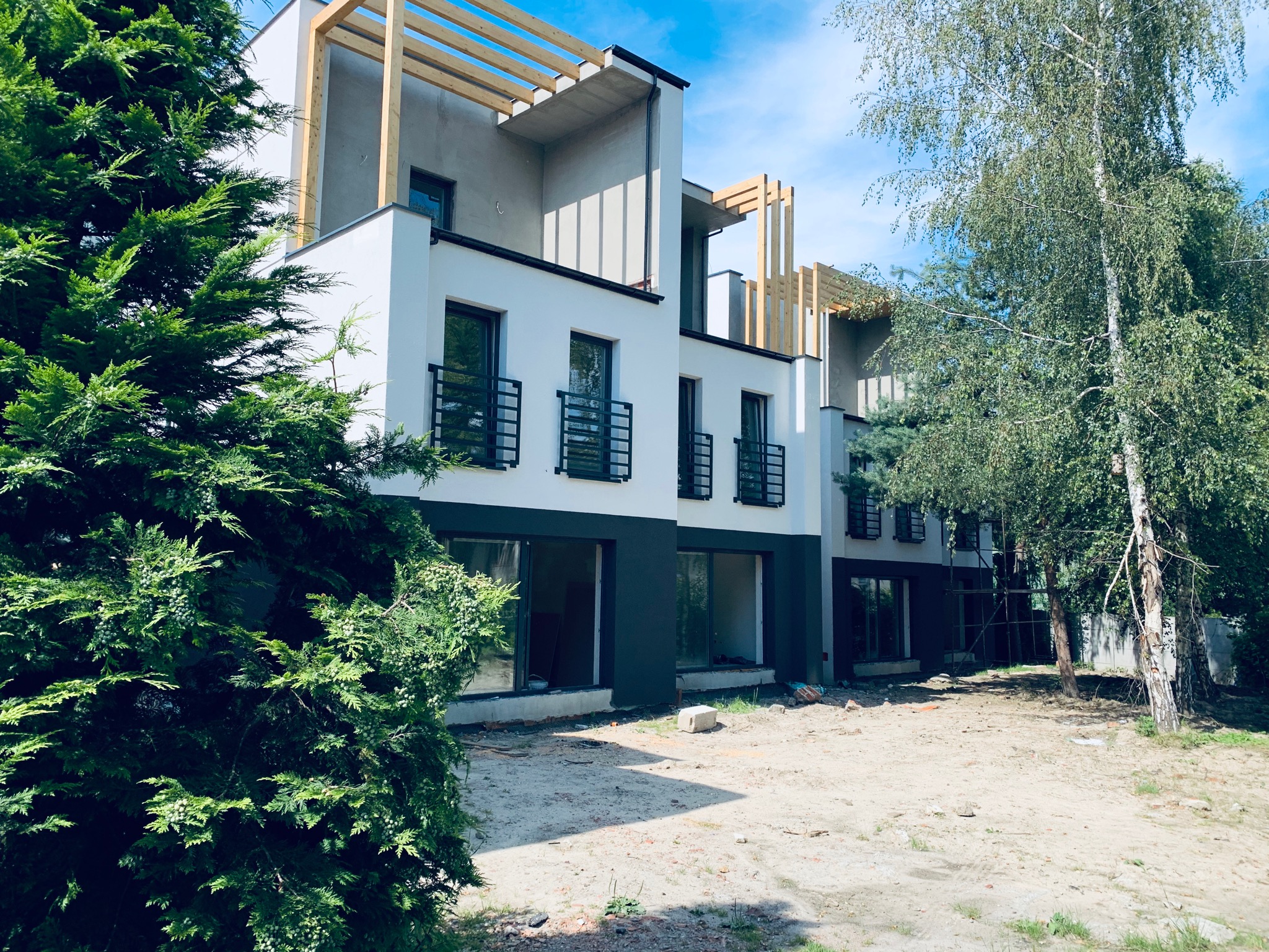 Nowoczesny budynek wielorodzinny w trakcie wykończenia elewacji, widoczne elementy konstrukcyjne drewniane, biała i grafitowa kolorystyka, surowy teren wokół budynku.