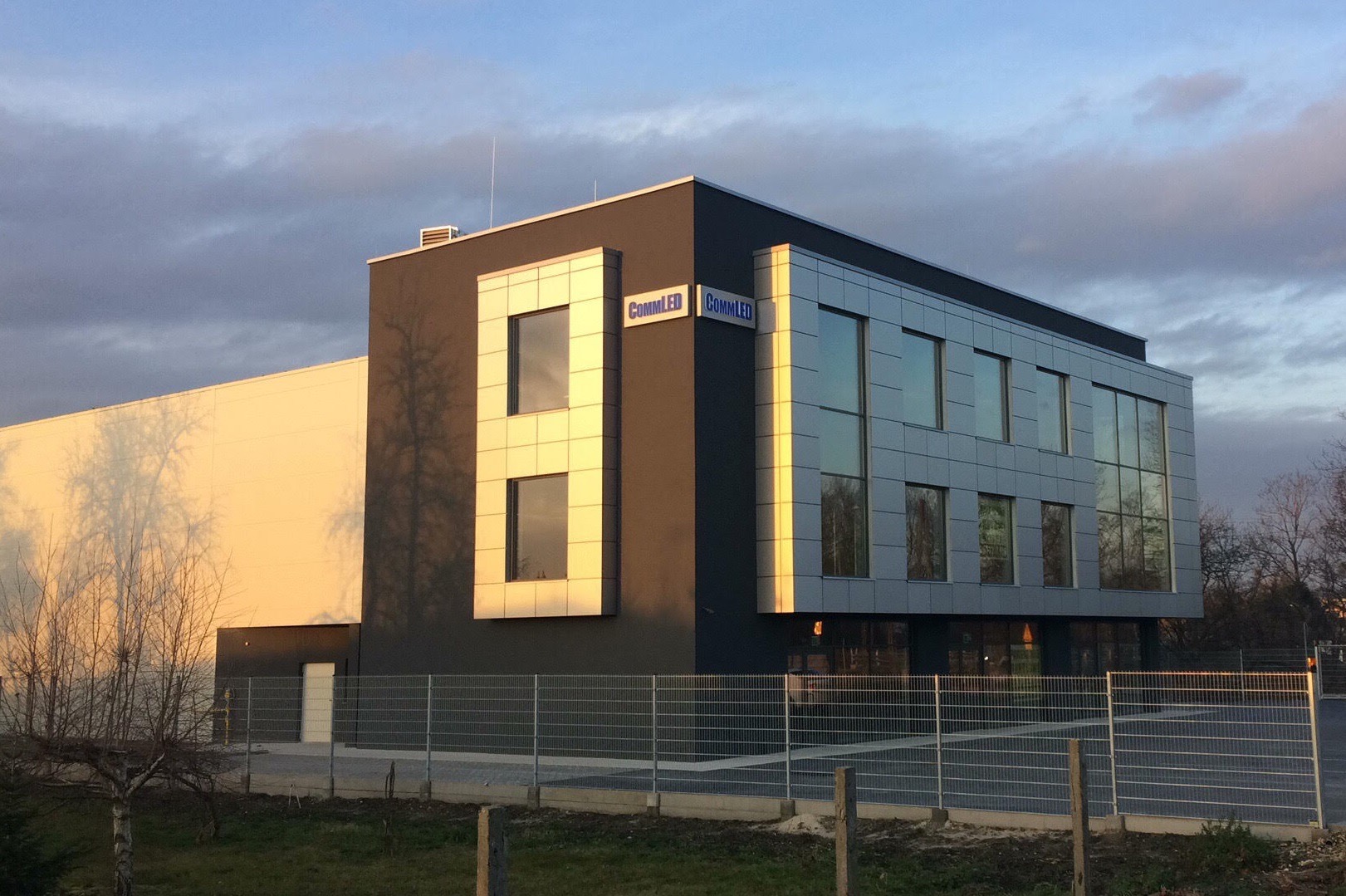 Nowoczesny budynek biurowy z szarą elewacją i aluminiowymi panelami, oświetlony popołudniowym słońcem, z widocznym logo firmy CommLED.
