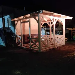 Niedokończona drewniana konstrukcja zadaszonej przestrzeni ogrodowej z dekoracyjnymi krzyżulcami, widoczna nocą na tle domu.