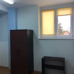 Pomalowane na jasno pomieszczenie biurowe z brązową szafą na dokumenty, niskim regałem i oknem z żółtymi roletami.