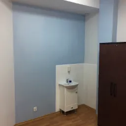 Świeżo pomalowana na niebiesko ściana w łazience, z umywalką w rogu i brązową szafką po prawej, widoczny fragment drewnianej podłogi i gniazdko elektryczne na ścianie.