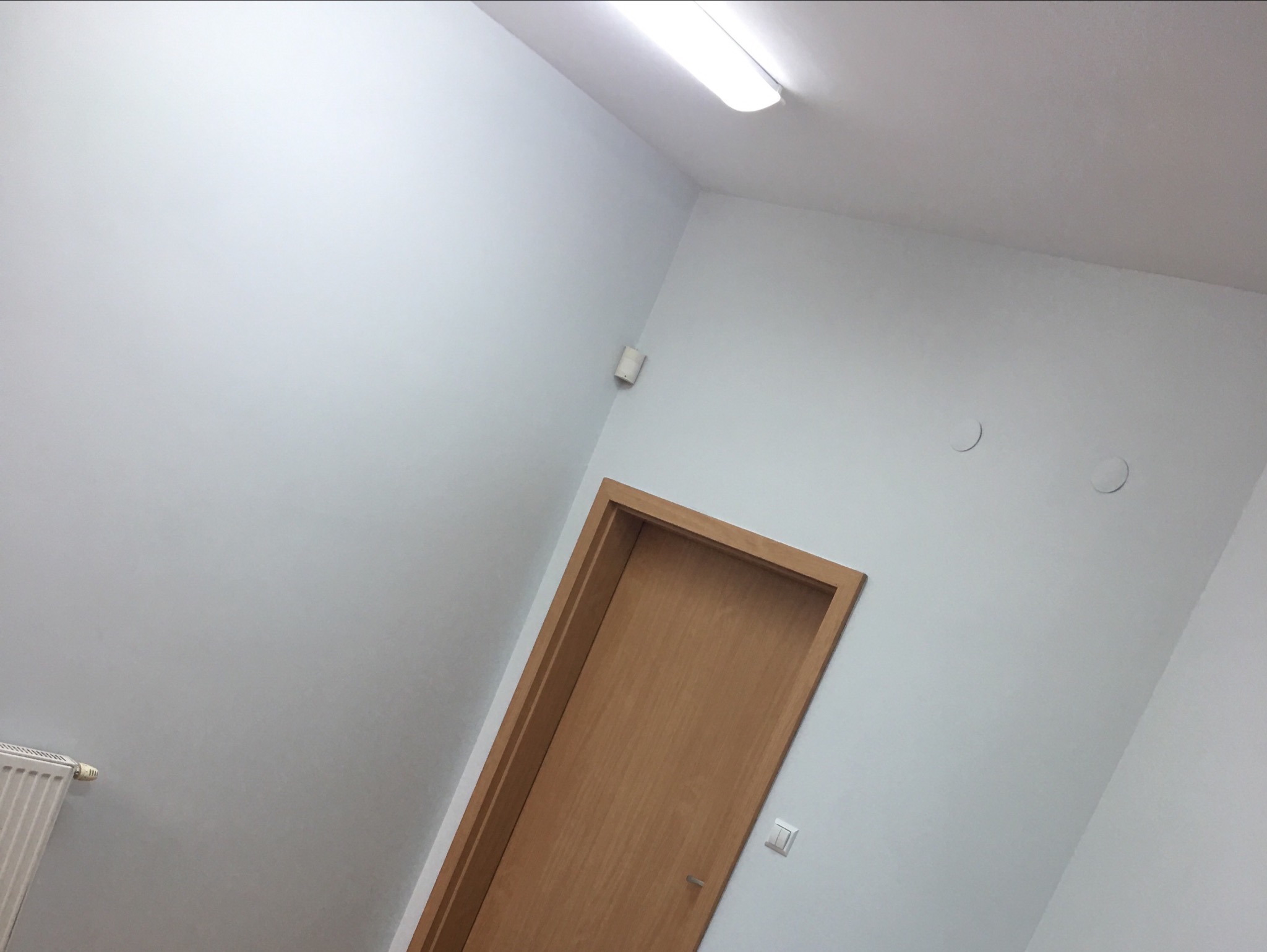 Drewniane drzwi wstawione w ościeżnicę, widoczne na tle świeżo pomalowanej, jasnoszarej ściany z widocznymi dwoma okrągłymi zaślepkami i wystającym grzejnikiem.