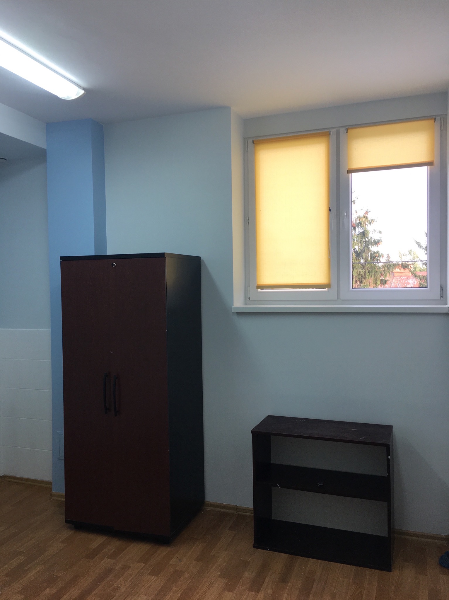 Pomalowane na jasno pomieszczenie biurowe z brązową szafą na dokumenty, niskim regałem i oknem z żółtymi roletami.