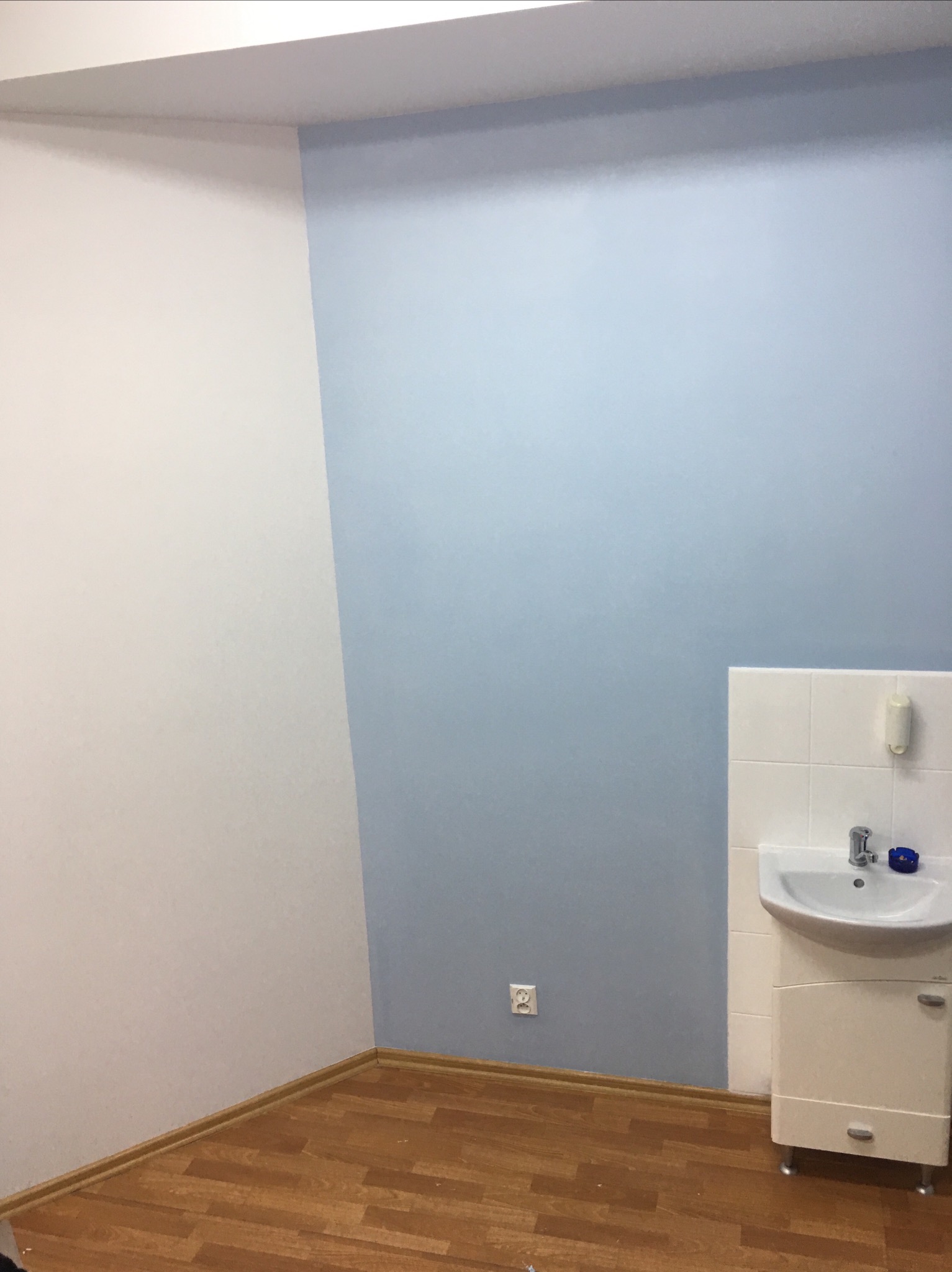 Pomalowany na dwa kolory róg pokoju z umywalką i szafką; ściana w kolorze jasnoniebieskim sąsiaduje z białą ścianą, podłoga wyłożona panelami imitującymi drewno.