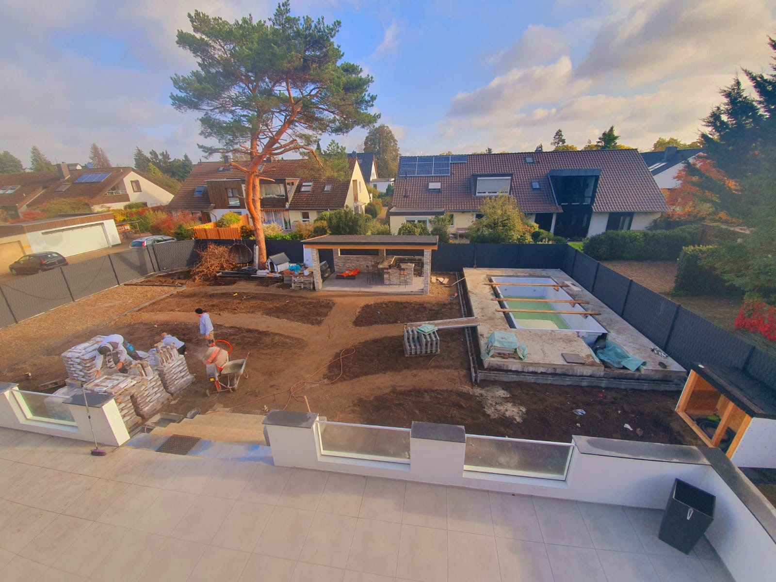 Wykonywanie prac ziemnych i budowlanych na posesji: przygotowanie terenu pod ścieżki, budowa altany z kamienia i fundamentu pod basen z zieloną wodą, widoczne worki z materiałami budowlanymi...