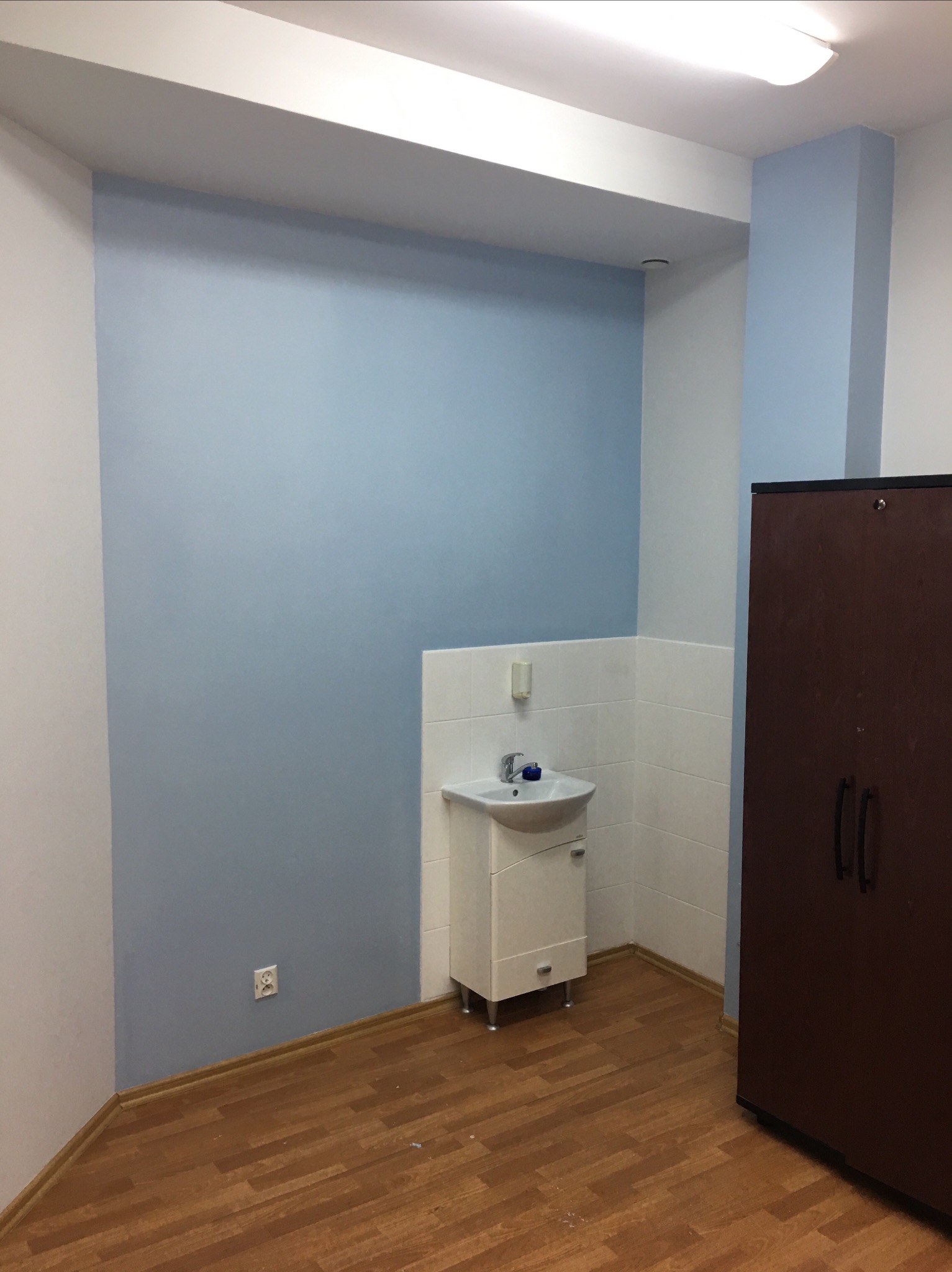 Świeżo pomalowana na niebiesko ściana w łazience, z umywalką w rogu i brązową szafką po prawej, widoczny fragment drewnianej podłogi i gniazdko elektryczne na ścianie.