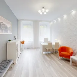 Jasne, przestronne biuro z białymi meblami, ceglaną ścianą i pomarańczowymi fotelami, obraz na ścianie, drewniana podłoga.