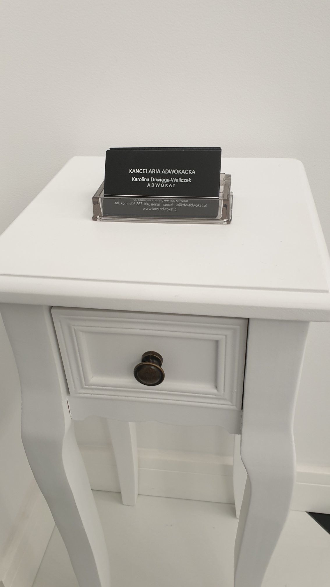 Wizytówka Kancelarii Adwokackiej Karoliny Drwięgi-Waliczek w przezroczystym stojaku na białym stoliku z szufladą.