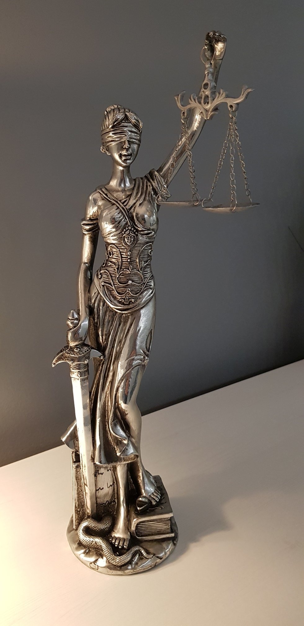 Metalowa statuetka Temidy z zasłoniętymi oczami, trzymająca wagę i miecz, stojąca na książkach i wężu.