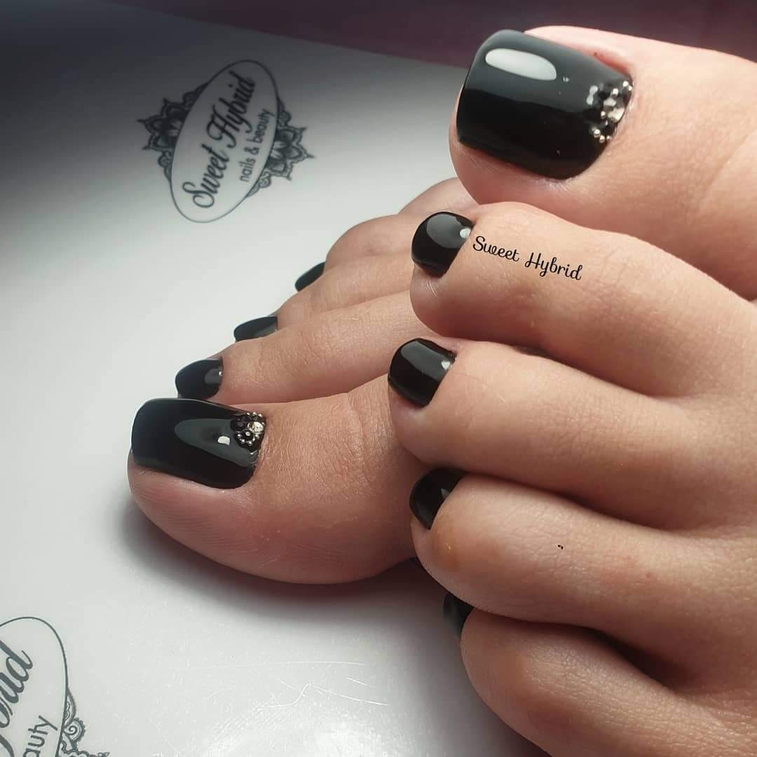 Pedicure w kolorze czarnym z delikatnymi zdobieniami na dużych palcach, wykonany lakierem hybrydowym marki Sweet Hybrid, widoczne logo firmy w tle.