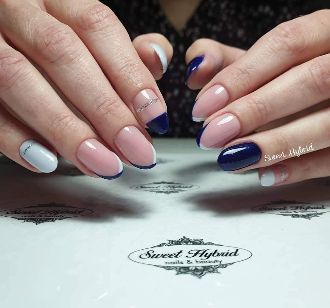 Dłonie prezentują manicure hybrydowy: połączenie klasycznego różu z granatowymi końcówkami i akcentem srebrnego paska na jednym paznokciu, na tle blatu z logo salonu kosmetycznego.