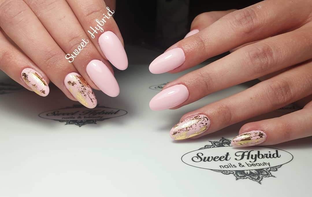 Delikatny, pastelowy róż na migdałowych paznokciach, akcentowany złotą folią na dwóch paznokciach każdej dłoni, na tle z logo salonu.