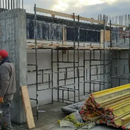 Wznoszenie ściany z białych bloczków, widoczne szalunki, stalowe zbrojenia, rusztowania i pracownik w czerwonej czapce, w tle zabudowa miejska.