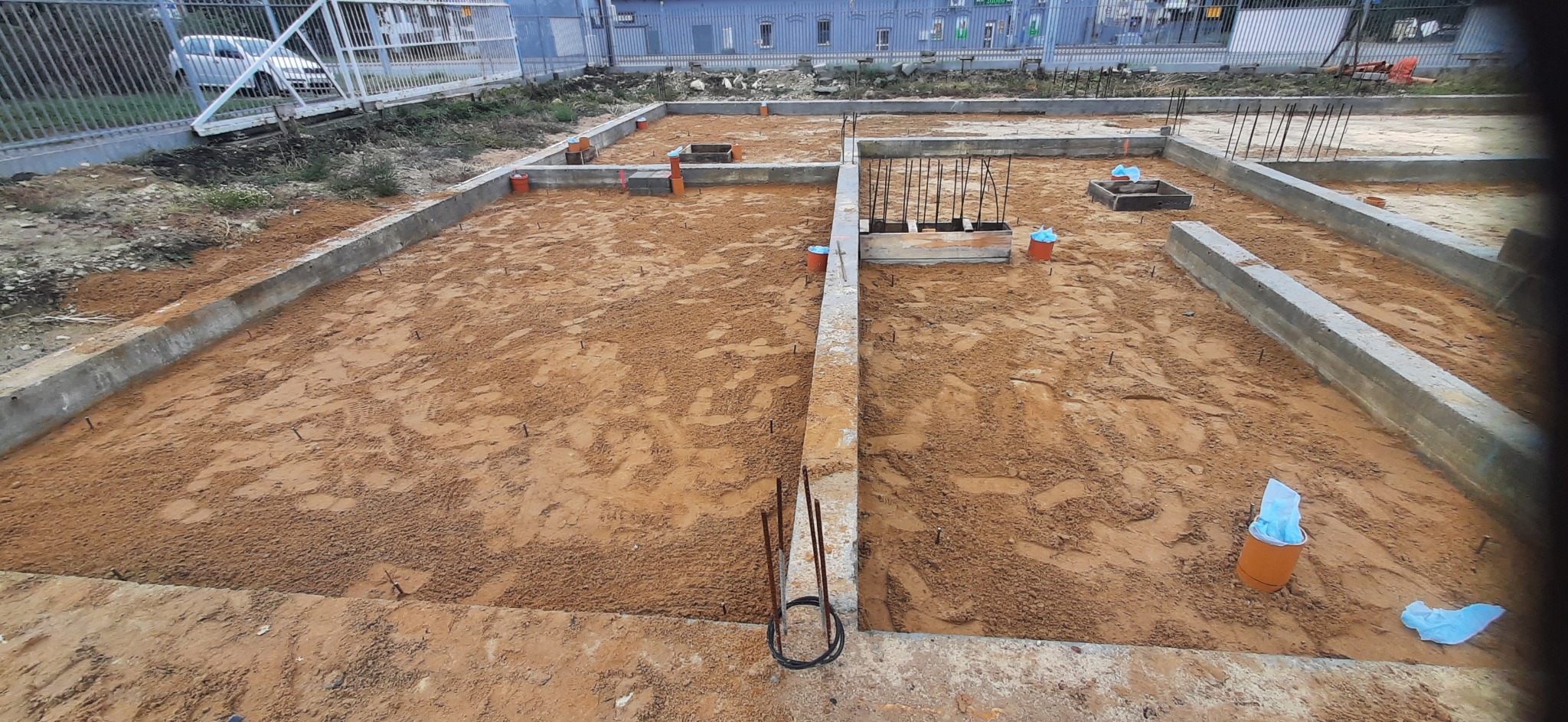 Wykop pod fundamenty z częściowo wylanymi betonowymi ścianami szalunkowymi, wystającymi prętami zbrojeniowymi i pomarańczowymi rurami kanalizacyjnymi, teren budowy zabezpieczony siatką.