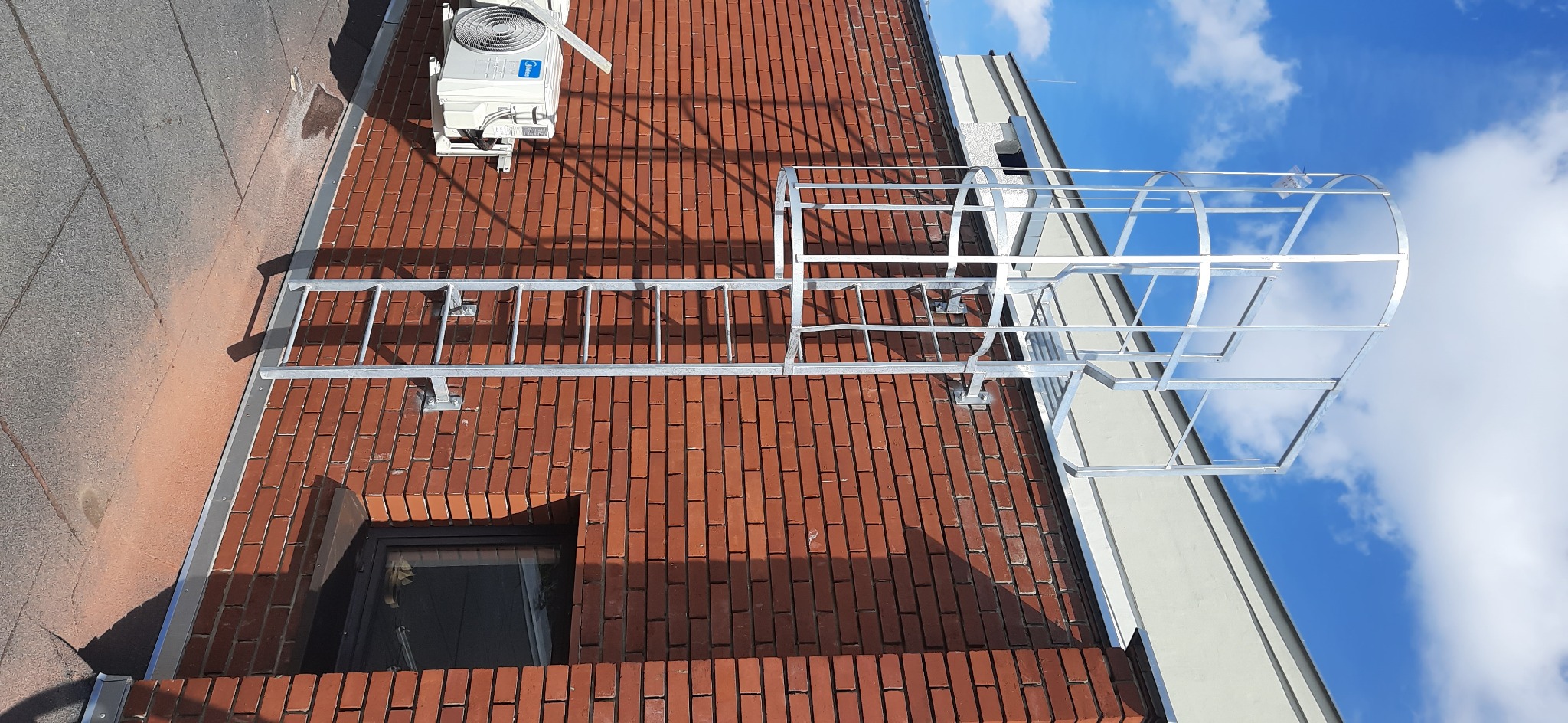 Zewnętrzna metalowa drabina ewakuacyjna z klatką bezpieczeństwa, zamocowana do ściany budynku z czerwonej cegły, widok z dołu na tle błękitnego nieba z chmurami.