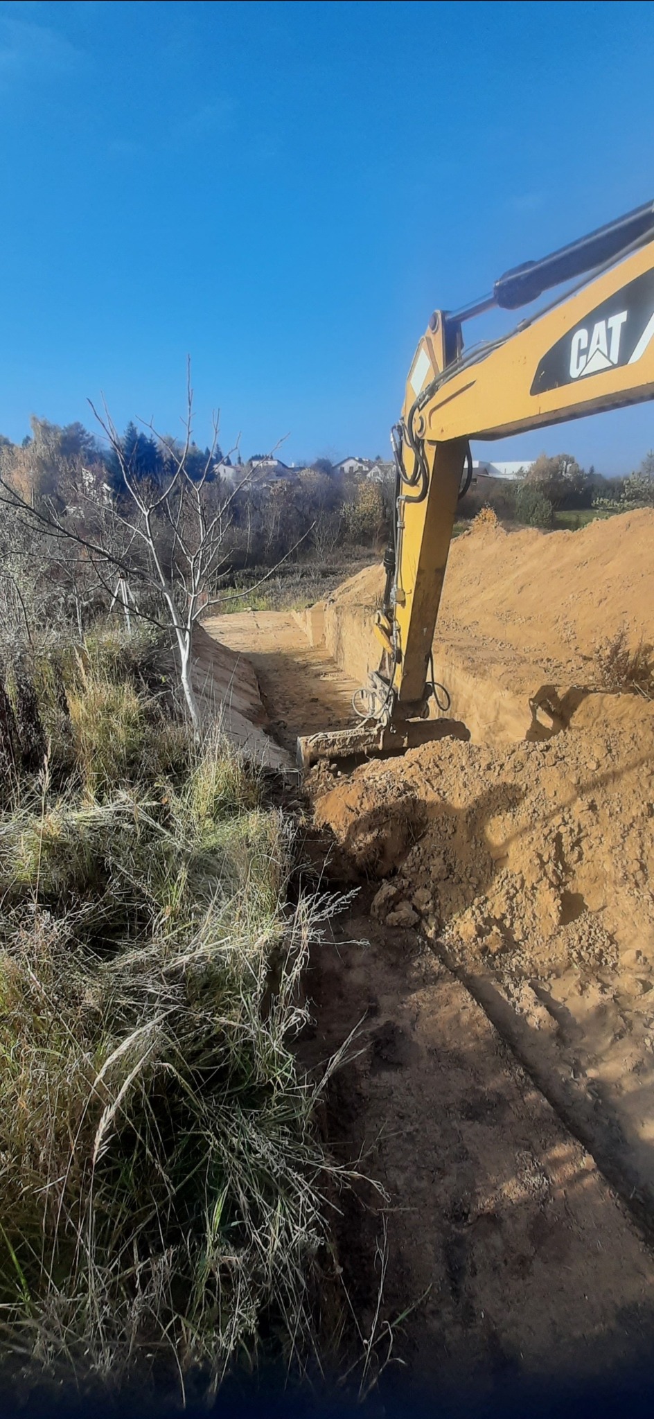 Żółta koparka CAT wykopuje rów w ziemi, widoczne zbocza wykopu i trawa na pierwszym planie, w tle zabudowania mieszkalne pod błękitnym niebem.