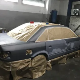 Samochód Audi 100 C4 przygotowany do lakierowania, z maskowaniem papierem i taśmą, widoczne szpachlowane fragmenty karoserii, w tle warsztat z wentylatorem i zbiornikiem ciśnieniowym.