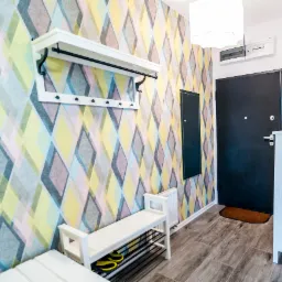 Przedpokój po renowacji z tapetą w romby, białą szafką na buty, wieszakiem z półką i czarnymi elementami, oraz ciemnymi panelami podłogowymi imitującymi drewno.