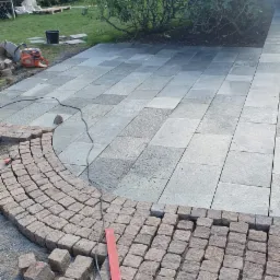 Fragment podjazdu z szarej kostki brukowej, w trakcie układania obrzeża z czerwonej kostki granitowej, widoczna poziomica i przecinarka do kamienia w tle.