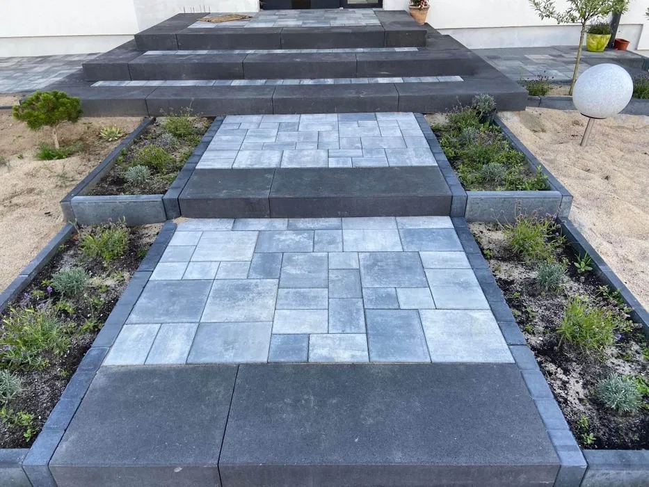 Nowoczesne wejście do domu z szarej kostki brukowej o nieregularnym wzorze, obrzeża z ciemnoszarego kamienia, niskie rabaty z roślinami po bokach, schody z ciemnoszarych bloków.