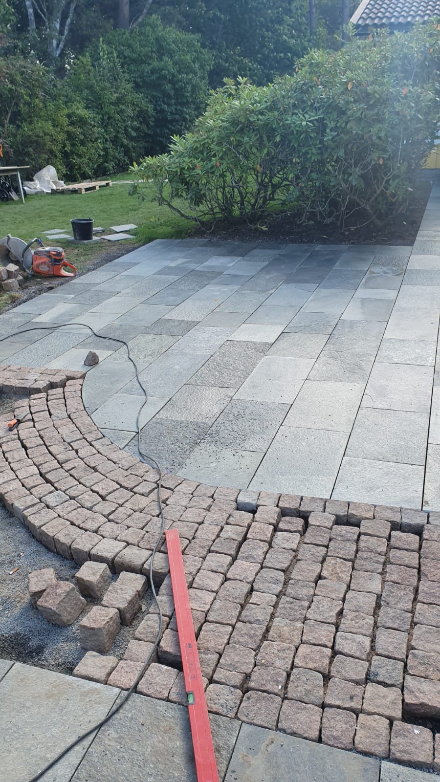 Fragment podjazdu z szarej kostki brukowej, w trakcie układania obrzeża z czerwonej kostki granitowej, widoczna poziomica i przecinarka do kamienia w tle.