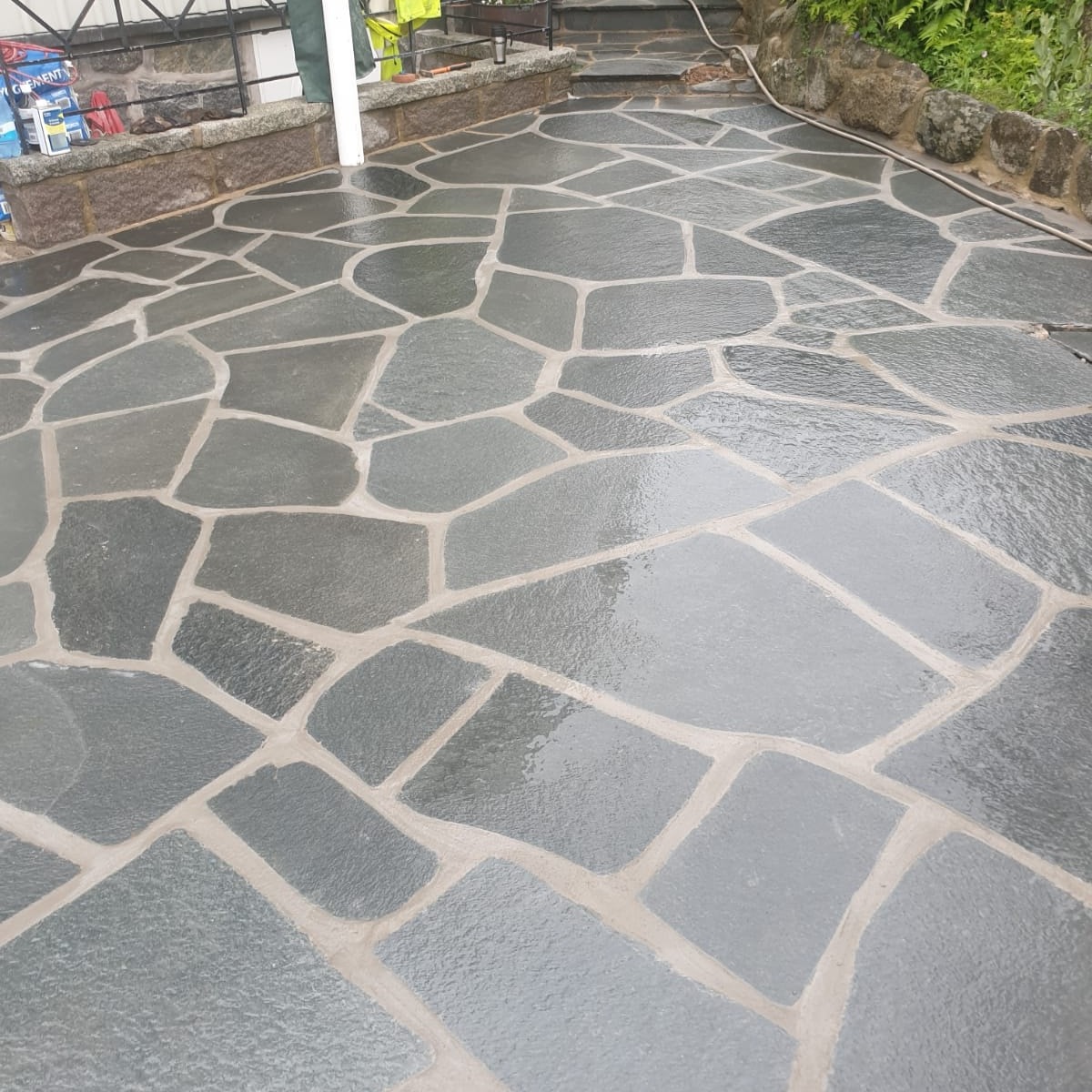 Nieregularna, szara kostka brukowa ułożona na posesji, z widocznymi fugami i mokrą powierzchnią, prowadząca do schodów i domu z elementami kamiennymi.