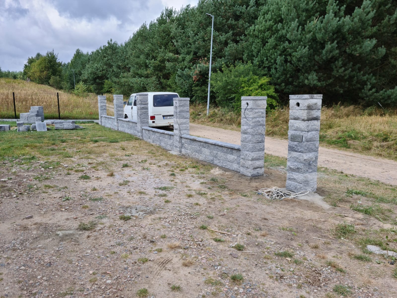 Nowo wybudowany, niski murek z szarej kostki łupanej, z filarami z tego samego materiału, częściowo ukończony, z widocznym stosem kostki obok i zaparkowanym białym vanem w tle, na działce...