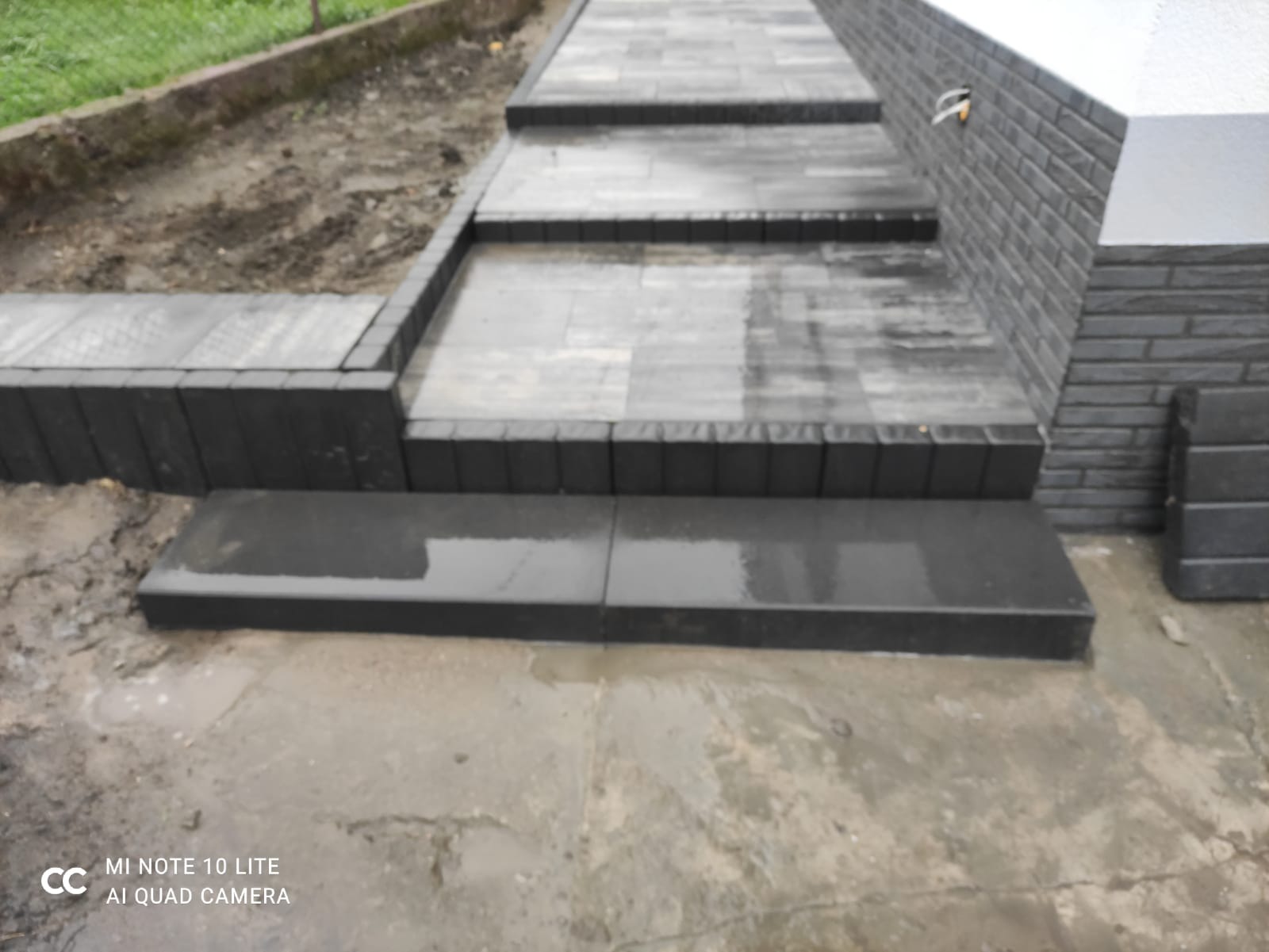 Schody zewnętrzne wyłożone ciemnoszarą kostką brukową, prowadzące w górę, po deszczu, z widocznymi refleksami wody na powierzchni, obrzeża z ciemnej kostki, obok ścieżka z ziemi.