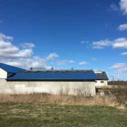 Budynek gospodarczy z niebieskim dachem i panelami słonecznymi na dachu, widok z poziomu trawy na tle błękitnego nieba z chmurami.