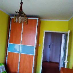 Wnętrze pokoju z żółtymi ścianami, szafą przesuwną z drewnianymi drzwiami i niebieskim pasem, oraz wiszącą lampą z motywem kwiatowym, widok na otwarte drzwi do kolejnego pomieszczenia.