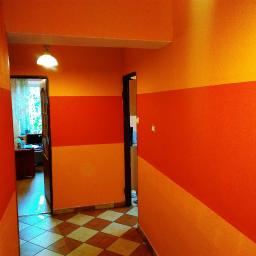 Bud-Om - Korytarz pomalowany w poziome pasy w odcieniach pomarańczy i żółci, z widoczną podłogą w szachownicę i otwartymi drzwiami do pomieszczeń po obu stronach.