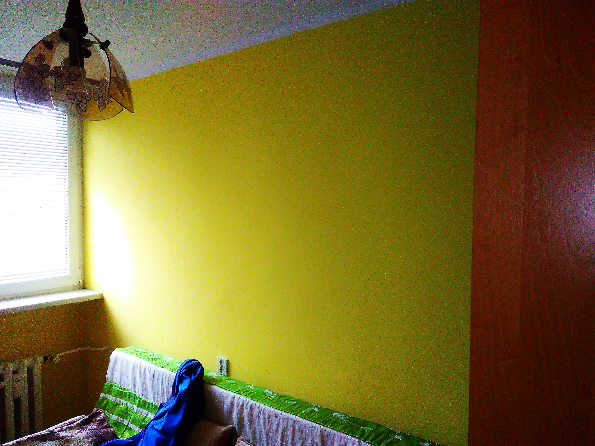 Ściana pomalowana na intensywny żółty kolor, widoczny fragment okna z żaluzjami, lampa z kwiatowym motywem i fragment szafy w odcieniach brązu. Na dole widoczny fragment mebla przykrytego tkaniną.