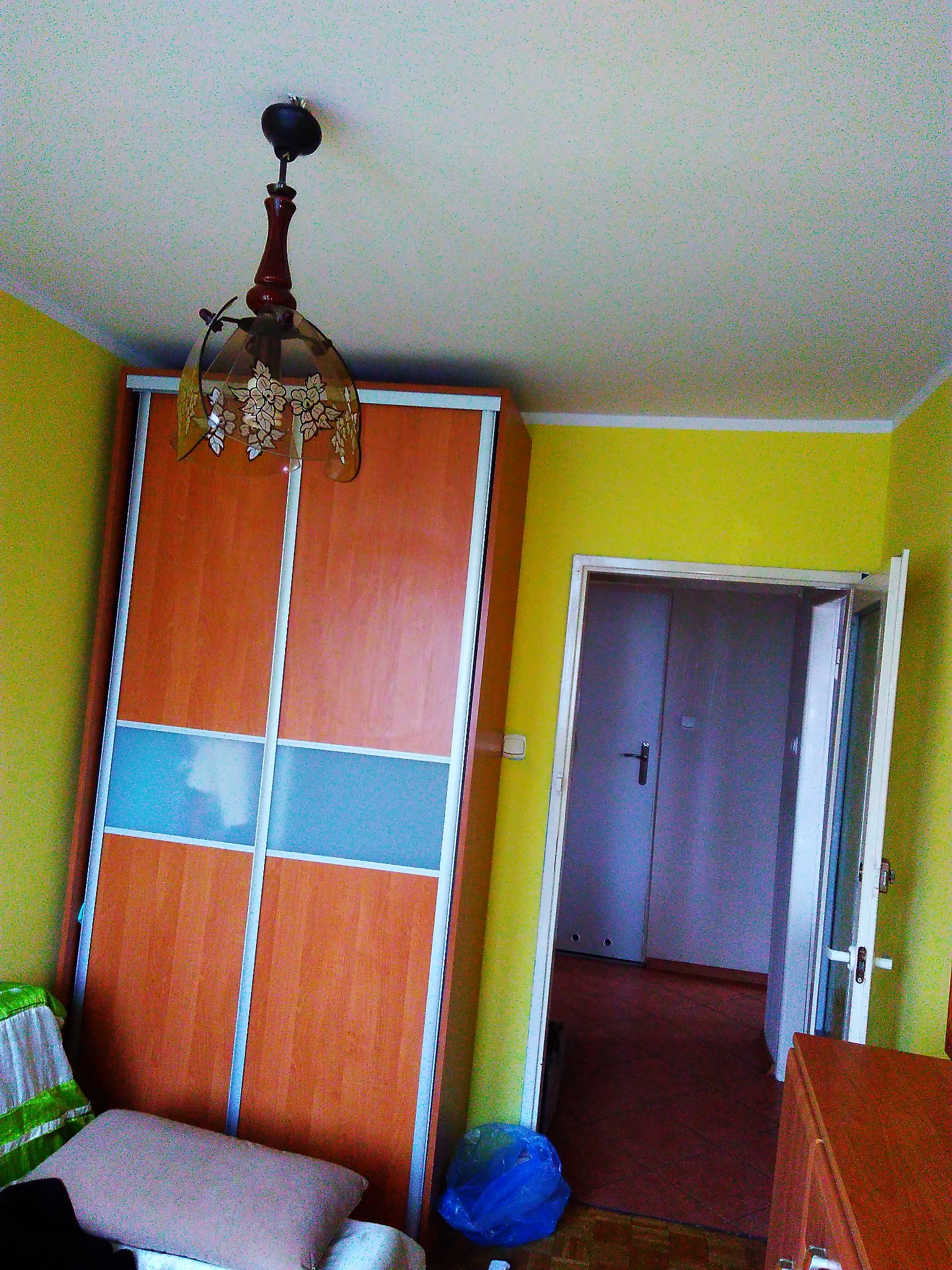 Wnętrze pokoju z żółtymi ścianami, szafą przesuwną z drewnianymi drzwiami i niebieskim pasem, oraz wiszącą lampą z motywem kwiatowym, widok na otwarte drzwi do kolejnego pomieszczenia.