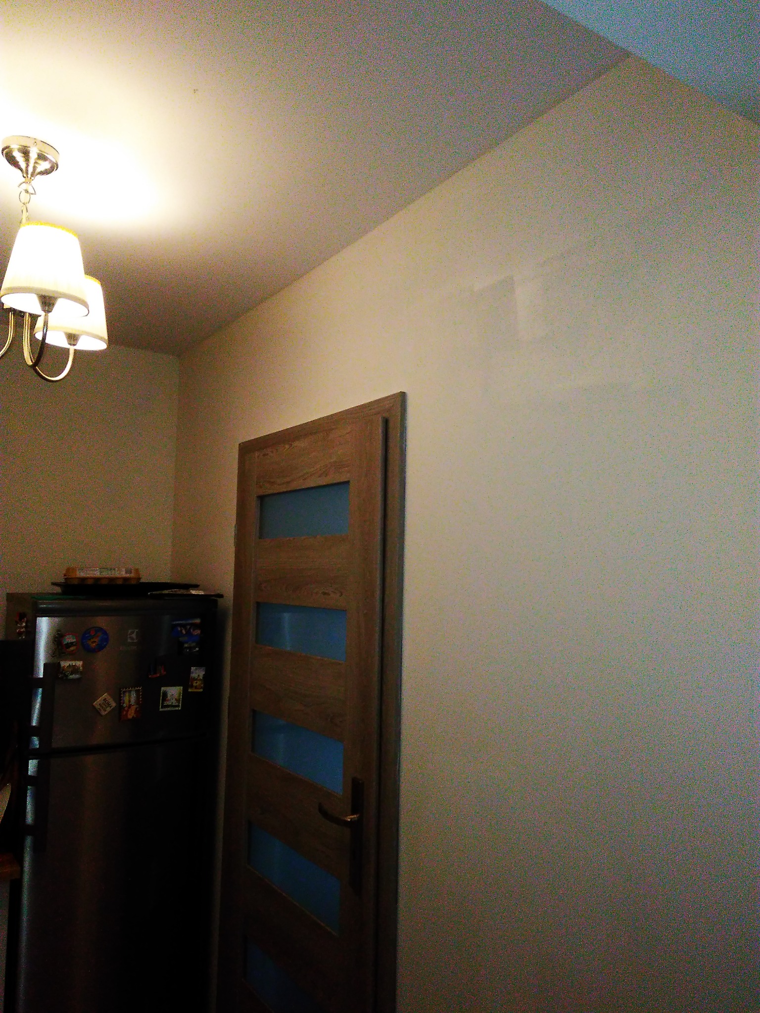 Ściana pomalowana na jasny kolor z widocznym śladem po naprawie, obok drewniane drzwi z przeszkleniami i lodówka z magnesami, oświetlona lampą sufitową.