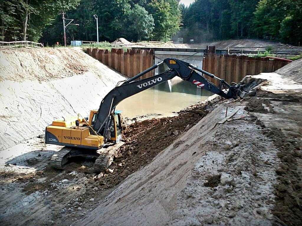 Żółta koparka Volvo na zboczu wykopu, z metalowymi ściankami zabezpieczającymi, pracująca przy budowie przepustu pod drogą.
