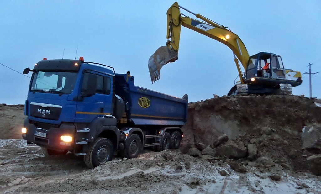 Żółta koparka Kobelco na wzniesieniu nabiera ziemię do niebieskiej wywrotki MAN z logo Meiller w błotnistym terenie.