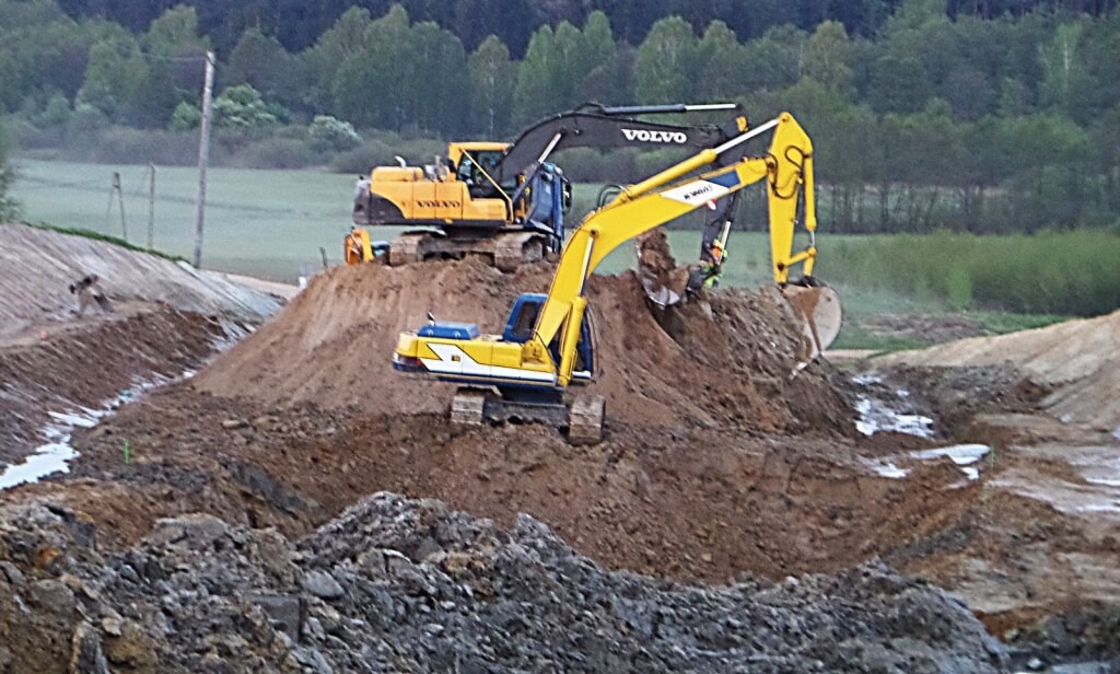Dwa żółte i czarne koparki Volvo na placu budowy, jedna kopie ziemię, w tle drzewa i pole, pochmurny dzień.