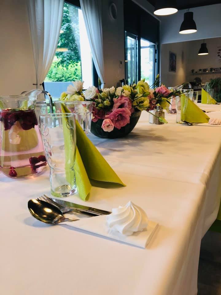 Elegancko nakryty stół z białym obrusem, szklankami, sztućcami, zielonymi serwetkami, dzbankiem z owocową wodą i dekoracją z kwiatów w ciemnej misie; na pierwszym planie beza na serwetce.