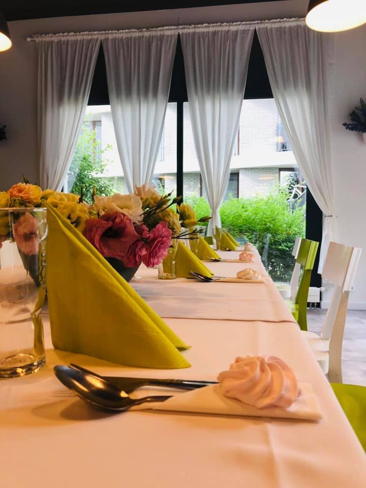 Elegancko nakryty stół z żółtymi serwetkami, kwiatami w wazonie i bezowymi babeczkami, ustawiony w jasnym pomieszczeniu z dużymi oknami i białymi zasłonami.