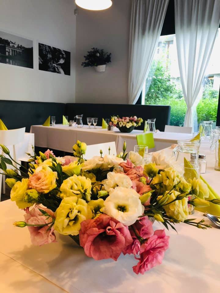 Uroczysto nakryty stół z bukietem różowych, żółtych i białych eustom na pierwszym planie, w tle zastawa stołowa, zielone serwetki i czarno-białe zdjęcia na ścianie.