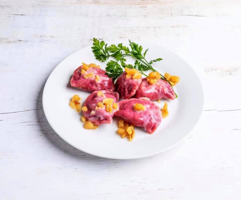 Porcja różowych pierogów z sosem i skwarkami, udekorowana gałązką zielonej pietruszki, podana na białym talerzu na tle białych desek.