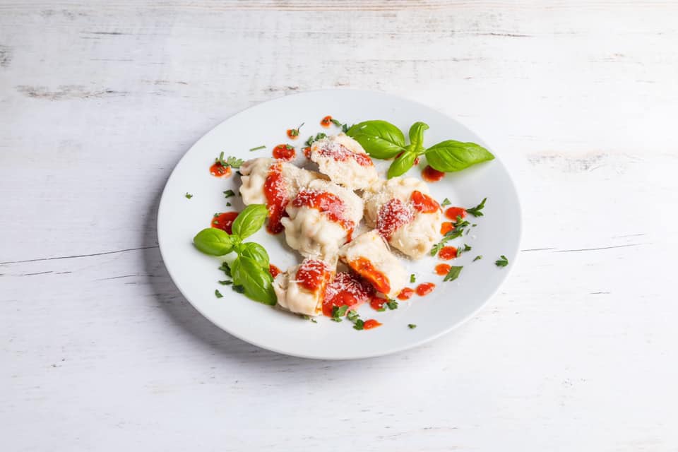 Delikatne pierogi tortellini z sosem pomidorowym i świeżą bazylią na białym talerzu, ułożone na jasnym, drewnianym stole.