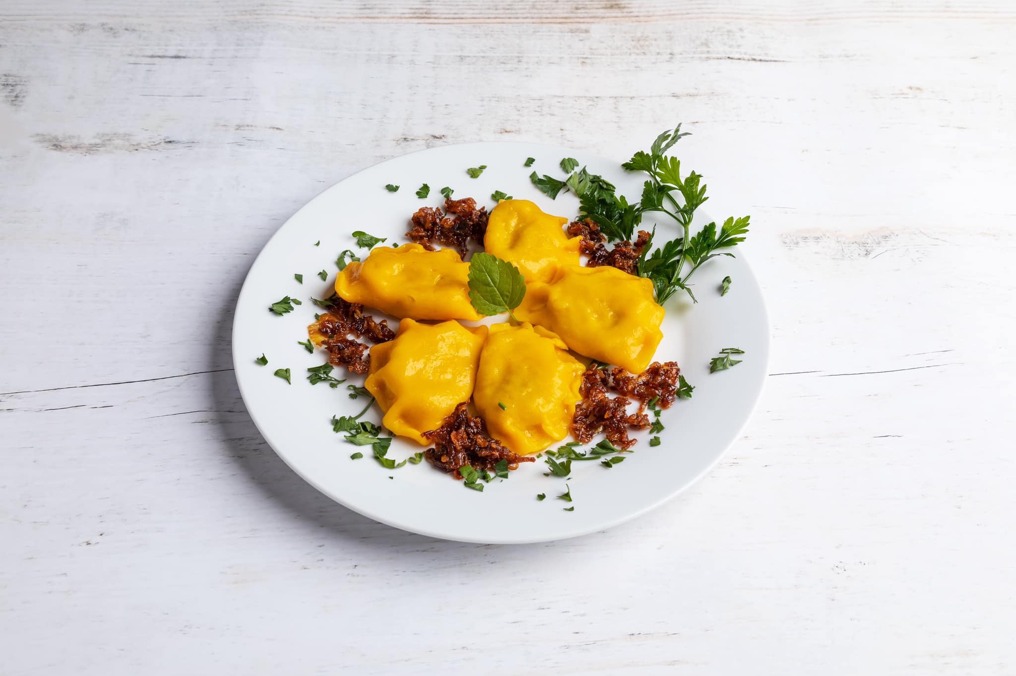 Pięć żółtych pierogów ułożonych na białym talerzu, udekorowanych karmelizowaną cebulą i świeżą pietruszką, całość na tle białych desek.