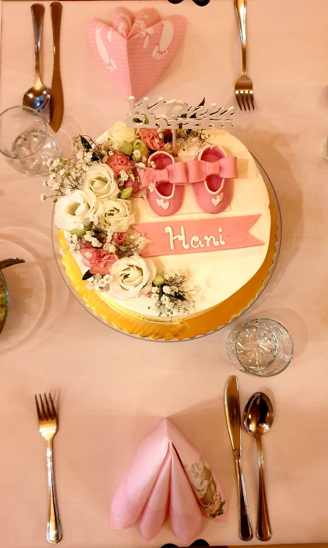 Udekorowany tort z napisem 'Hani', różowymi baletkami z lukru i kwiatami, otoczony nakryciem stołu z różowymi serwetkami i złotymi sztućcami, na jasnoróżowym obrusie, sugerujący przyjęcie z okazji...