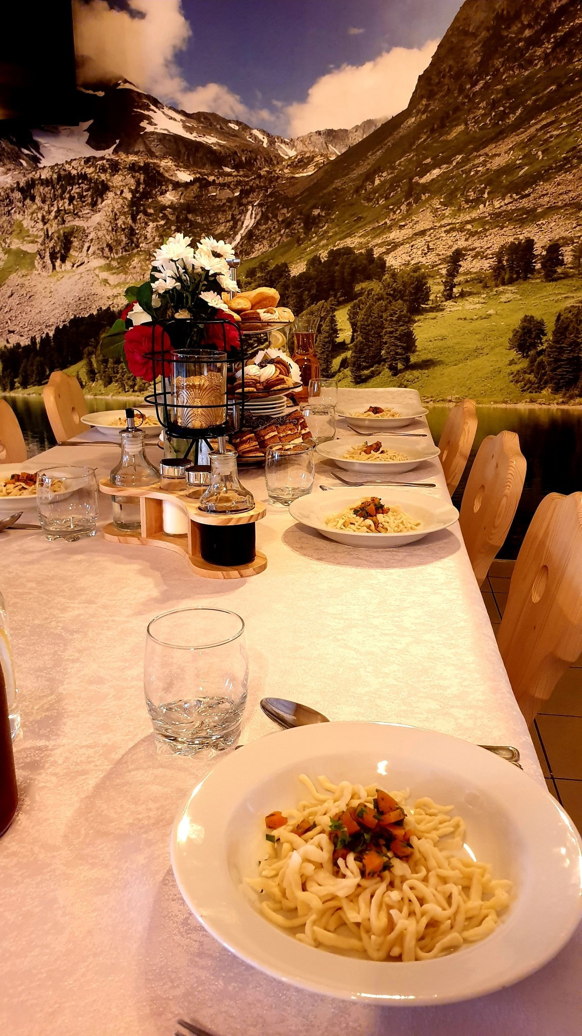 Uroczysty stół w Nowym Sączu, nakryty białym obrusem z talerzami z kluskami i sosem, szklankami, oraz dekoracjami kwiatowymi i deserami, z fototapetą górską w tle.
